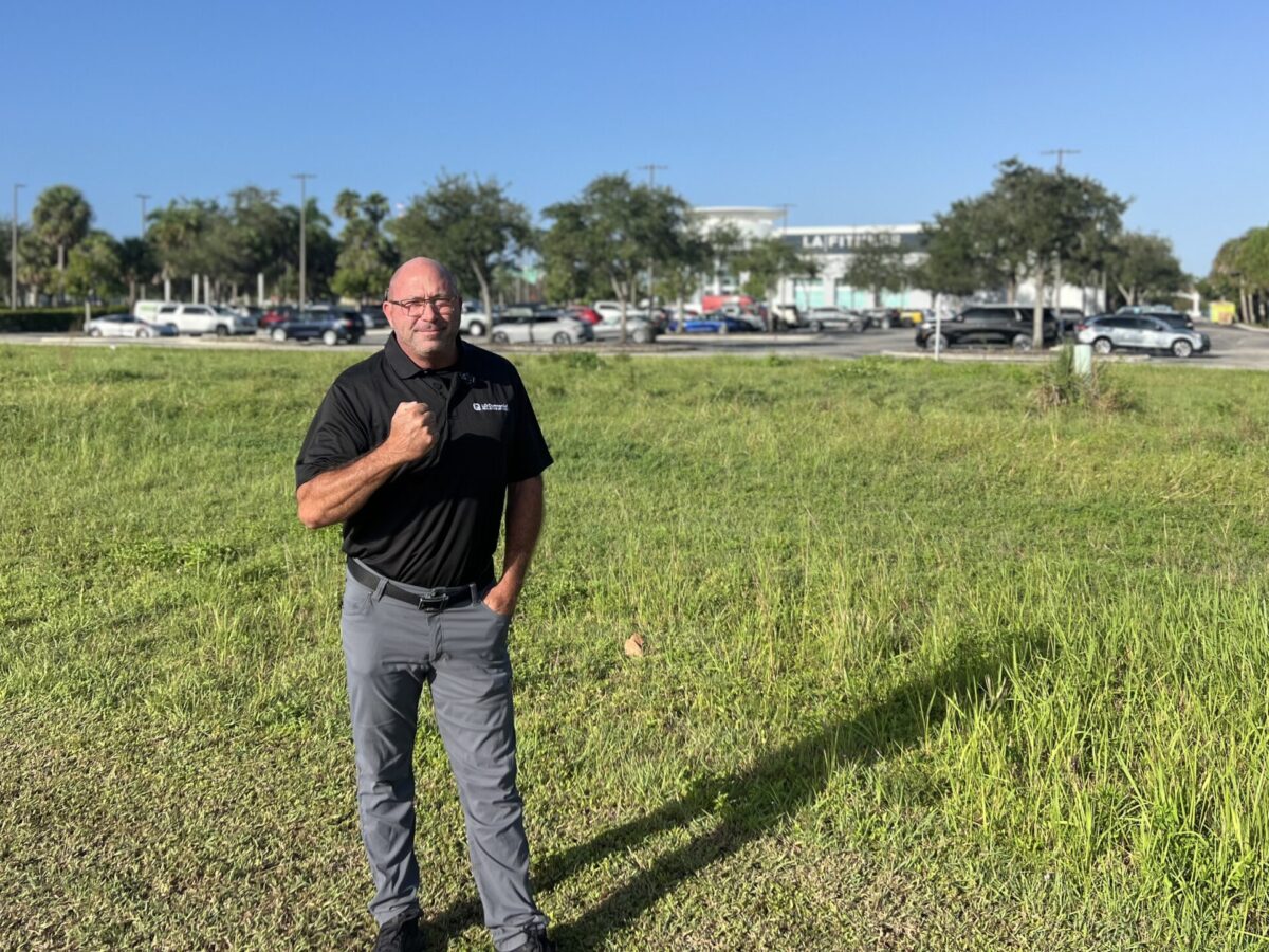Man standing in grassy field, parking lot background.