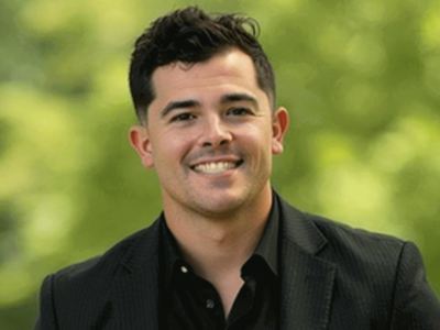 Smiling person in a black suit outdoors.